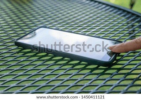 Person's finger tapping on touch button using smart cell phone. Modern tech outside resting on textured pattern outdoor patio table. Sun reflecting glare off screen. Communication in information age.