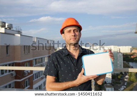 builder in a protective helmet with a tablet for writing in his hand is standing on the roof of a building overlooking the buildings Construction architecture business engineer at a construction site