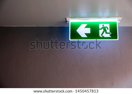 Fire exit sign with icon and arrow pointing to the left on green background hanging from the ceiling.