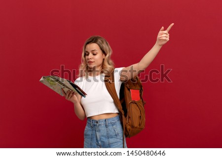 Blond tourist with curly hair in a white t-shirt pointing with a hand at the right during sightseeing tour holding map and orange backpack. Concept of travel