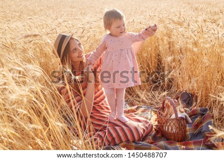 Outdoor shot of beautiful woman with her little doughter toddler wearing pink dress and tights in wheat field on summer day, mommy dressed striped dress and straw hat posing surrounded with spikelets.
