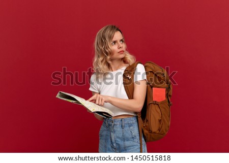 Confused blond girl with curly hair in a white t-shirt trying to find itinerary with the map holding brown orange backpack. Girl lost at the unknown place. Concept of travel Royalty-Free Stock Photo #1450515818