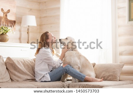 Young pretty woman in casual clothes hugging her beloved big white dog sitting on the sofa in the living room of her cozy country house. Animal communication concept Royalty-Free Stock Photo #1450573703