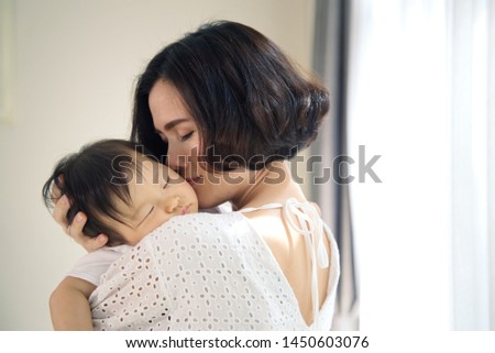 Asian beautiful mother hugging sleeping baby in her arms and kissing the kid gently. The mom closing her eyes while holding her baby head to rest on shoulder. Touch of love and family relationship.