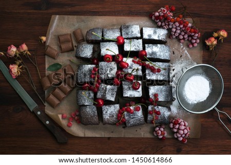 Homemade brownie (cake) on parchment paper. Near cherry, icing sugar and red currants. Brown wood background