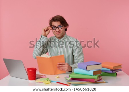 Young cheerful man in glasses wears in shirt, sits by the table and reads book, looks at the camera through glasses, working with notebook, prepared for exam, isolated over pink background.