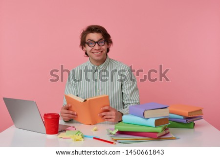 Young cheerful man in glasses wears in shirt, sits by the table and working with notebook, prepared for exam, reads book, looks happy and enjoys the reading, isolated over pink background.