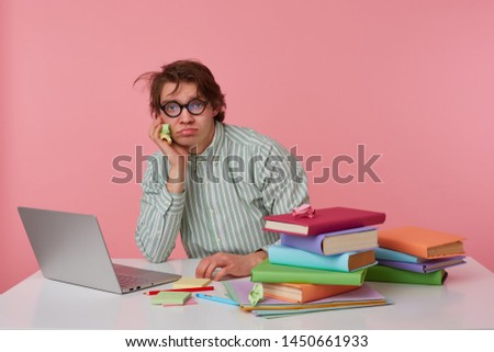 Young displeased guy with glasses, wears on blank shirt, sitting at a table with books, working at a laptop, looks unhappy and bored, sadly looks at the camera isolated over pink background.