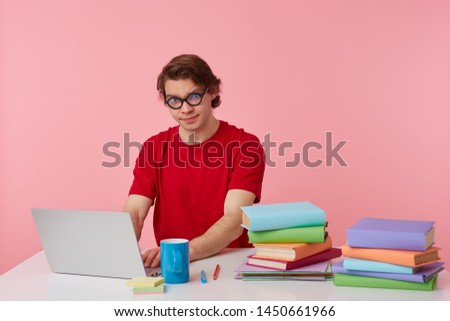 Photo of young student in glasses wears in red t-shirt, man sits by the table and working with laptop and books, isolated over pink background. Looks displeased and unhappy.