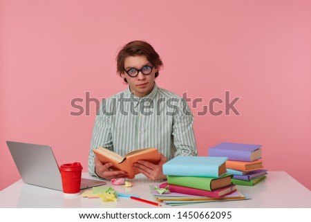 Photo of young man in glasses wears in shirt, student sits by the table and working with notebook, prepared for exam, reads book, having serious look, isolated over pink background.
