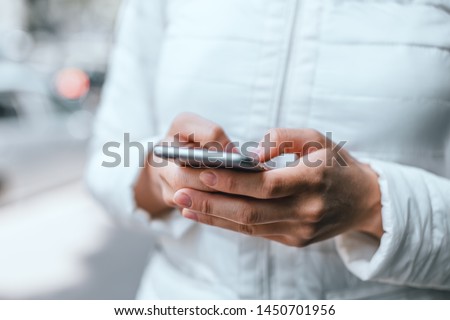 Close up of woman's hands in white jacket holding grey smartphone and typing a message with both hands on street. The side of the phone. Royalty-Free Stock Photo #1450701956