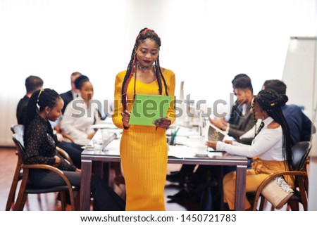 Face of handsome african business woman in yellow dress, holding clipboard on the background of business peoples multiracial team meeting, sitting in office table.