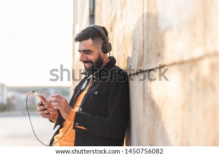 Hipster Latin Man Listening Music on Headphones Outdoors - Handsome Latin Guy Feeling the Music in the City - Latin American Guy Using Cell Phone and Listening Music. Lifestyle Concept.