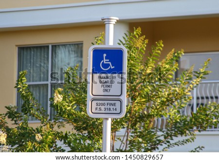 A sign with the wheelchair logo stating: "Parking only with disability permission", $ 200 fine. In the background a cream-colored house with a bush.