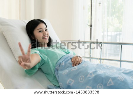 Young Asian patient woman lying at hospital bed with saline drip showing victory sign for cheerful. Drop of saline solution to help patient. Healthcare and health lifestyle, medical concept.