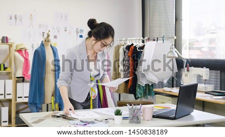 elegant female designer working on laptop and sketching at desk in studio. young woman fashion worker in workshop looking examining at paper of new collection checking with mannequin standing behind