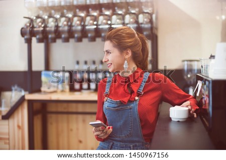 pretty young trendy woman wearing red shirt and denim casual overalls smiling in coffee house with cellphone in hand