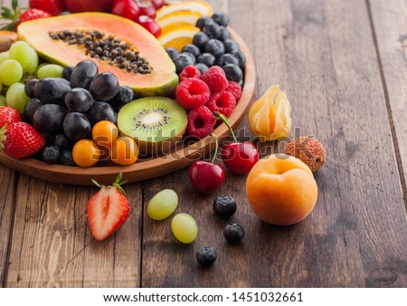 Fresh raw organic summer berries and exotic fruits in round wooden plate on wooden kitchen background. Papaya, grapes, nectarine, orange, raspberry, kiwi, strawberry, lychees, cherry Top view Royalty-Free Stock Photo #1451032661