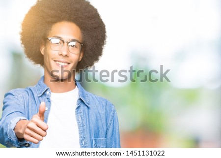 Young african american man with afro hair wearing glasses doing happy thumbs up gesture with hand. Approving expression looking at the camera with showing success.