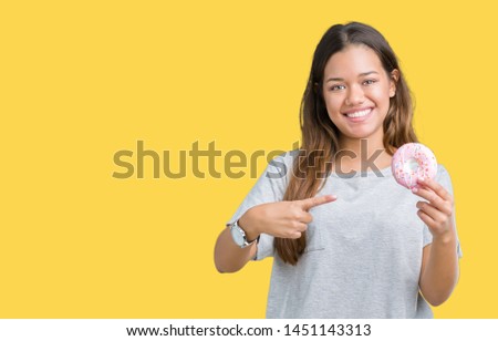 Young beautiful woman eating pink donut over isolated background very happy pointing with hand and finger