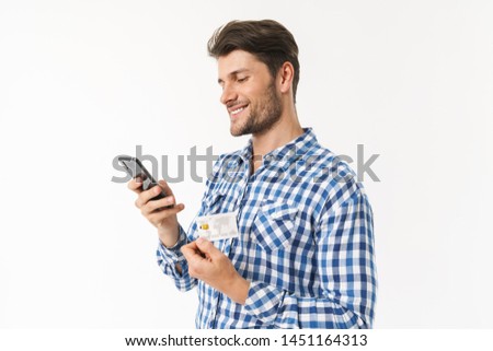 Photo of happy smiling handsome young man standing isolated over white wall background dressed in casual shirt using mobile phone holding credit card.