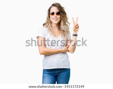 Beautiful young blonde woman wearing sunglasses over isolated background smiling with happy face winking at the camera doing victory sign. Number two.