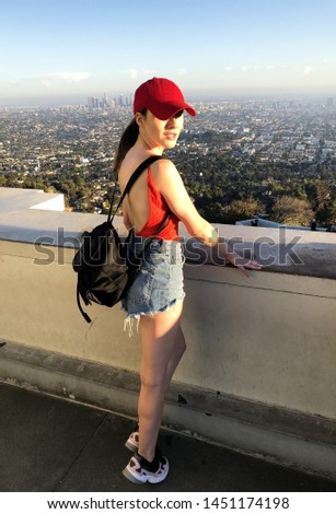 Model posing on the background of Los Angeles, from Griffith Observatory.  Dressed in red top, blue shorts,  black backpack, denim sports shoes and a red baseball cap.  Photo taken on a sunny day