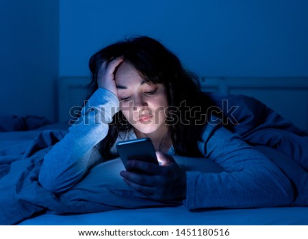 Addicted young woman chatting and surfing on the internet using her smart phone sleepy, bored and tired late at night. Dramatic dark light. In Internet, Mobile addiction and insomnia concept. Royalty-Free Stock Photo #1451180516