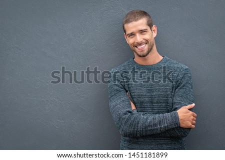 Portrait of happy young man leaning against wall isolated on grey background with big smile. Cheerful guy in winter clothes on gray wall looking at camera. Happy man wearing sweater with copy space.