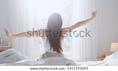 Beautiful Brunette is Waking up in the Morning, Stretches in the Bed, Sun Shines on Her From the Big Window. Happy Young Girl Greets New Day. Royalty-Free Stock Photo #1451195093
