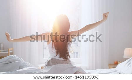 Beautiful Brunette is Waking up in the Morning, Stretches in the Bed, Sun Shines on Her From the Big Window. Happy Young Girl Greets New Day with Warm Sunlight Flare. Royalty-Free Stock Photo #1451195096