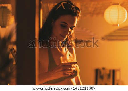 Young beautiful woman at the beach bar kiosk having a drink at sunset, summertime and vacations concept