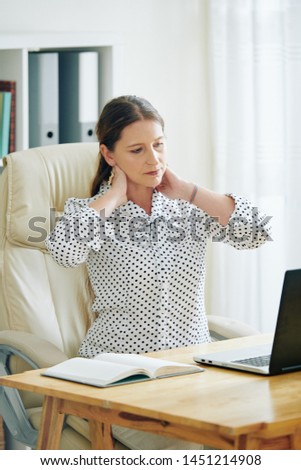 Woman rubbing stiff neck when sitting at office desk and reading e-mail or contract on laptop