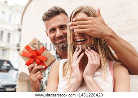 Image of charming excited couple in summer clothes smiling and holding present box together while standing against wall on city street