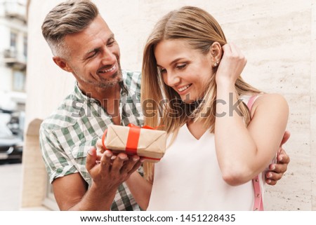 Image of amazed excited couple in summer clothes smiling and holding present box together while standing against wall on city street