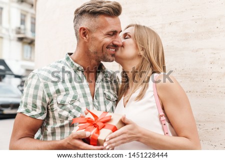 Image of pretty excited couple in summer clothes kissing and holding present box together while standing against wall on city street