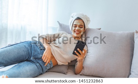 Young woman with white clay facial mask using smartphone while sitting on bed at home. Woman having face treatment at home lying on grey bed. Woman use beauty mask with cellphone at home