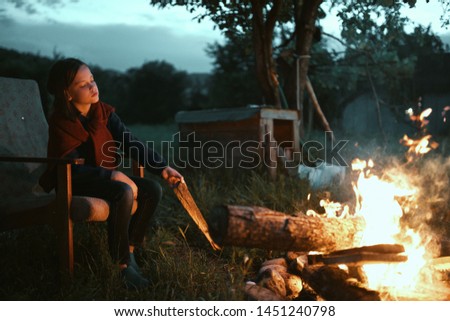 A pretty girl in nature sits throwing wooden shelves into a fire and an ancient armchair an evening of rest