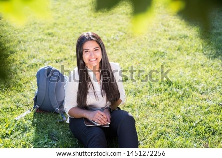 Indian girl, student, sitting on the green grass, with a backpack, smiling and holding a tablet