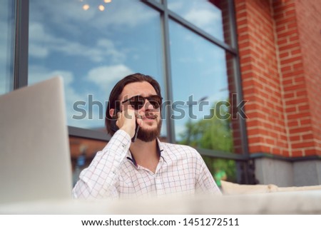 Photo of men in plaid shirt talking on phone while sitting at table with laptop near brick building