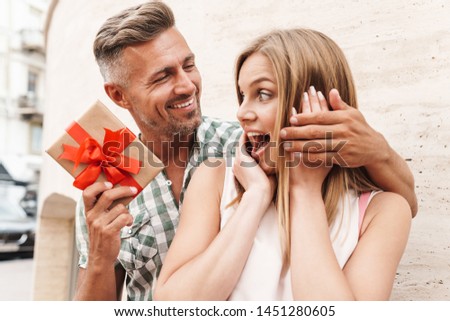 Image of astonished excited couple in summer clothes smiling and holding present box together while standing against wall on city street