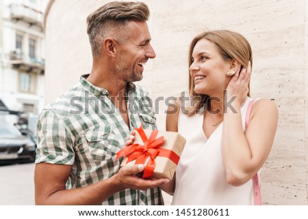 Image of romantic excited couple in summer clothes smiling and holding present box together while standing against wall on city street