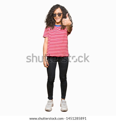 Young beautiful woman with curly hair wearing sunglasses doing happy thumbs up gesture with hand. Approving expression looking at the camera with showing success.