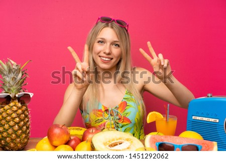 Young blonde woman with lots of fruits smiling and showing victory sign