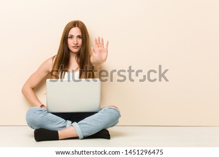 Young ginger woman sitting on her house floor standing with outstretched hand showing stop sign, preventing you.