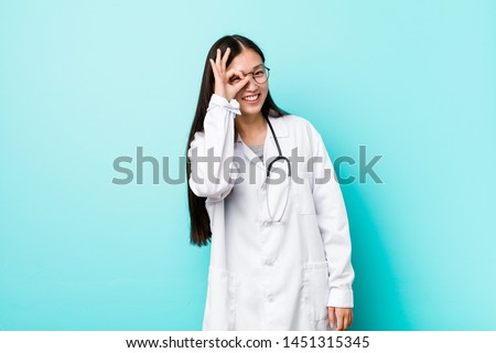 Young chinese doctor woman excited keeping ok gesture on eye.