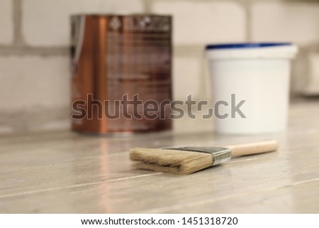 A paint brush is next to a plastic paint bucket with a blue lid and metal can on an old white vintage wooden board. Brick wall in the background. Place for text or logo.