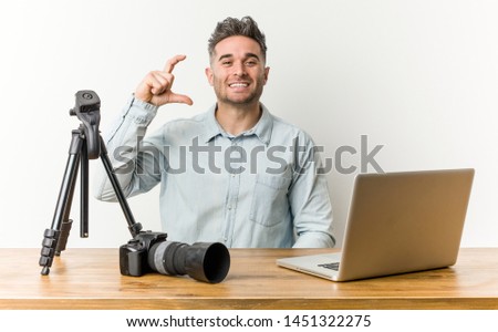 Young handsome photography teacher holding something little with forefingers, smiling and confident.