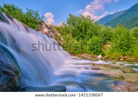 A fast cold turquoise river runs along trees and stones in the north of Italy in the Alps. The picture was taken on a long exposure.
