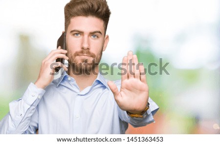 Young handsome man business talking on smartphone with open hand doing stop sign with serious and confident expression, defense gesture